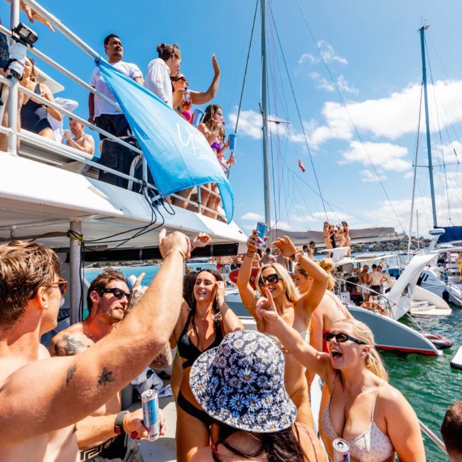 Group of people partying on a boat under clear skies, with some raising drinks and others dancing. Multiple boats are visible in the background on the water at a luxury yacht hire in Sydney.