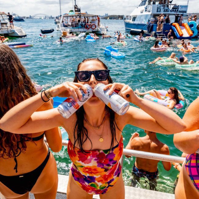 People on boats and in water enjoying a lively party on a sunny day. A woman in the foreground is drinking from two cans simultaneously. This Catamaran party in Sydney features vibrant music, thanks to DJ boat hire, creating an unforgettable experience under the sun.