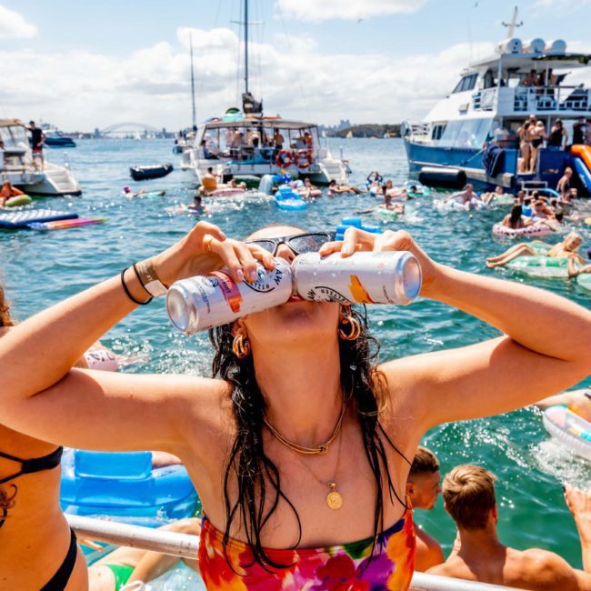 A person on a catamaran party in Sydney is drinking from two cans simultaneously. Other boats and people swimming and floating are visible in the background. The scene is lively and sunny.