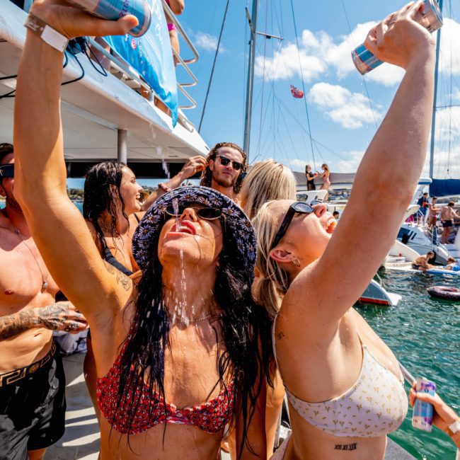 Two women in bikinis pour drinks over themselves while standing on a private yacht charter Sydney Harbour, surrounded by people, with a banner in the background and sailboats visible in the water.