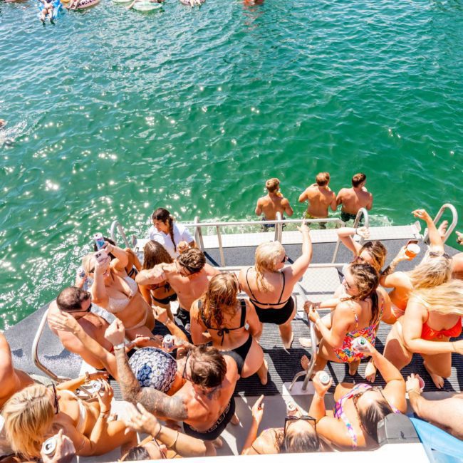 A large group of people on a catamaran party in Sydney, wearing swimsuits and enjoying a sunny day, with several individuals swimming in the water nearby.