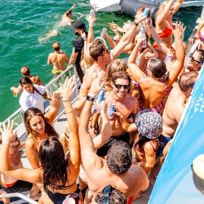 A group of people in swimsuits celebrate and socialize on a luxury yacht near Sydney, some holding drinks, with a swimmer approaching the boat.