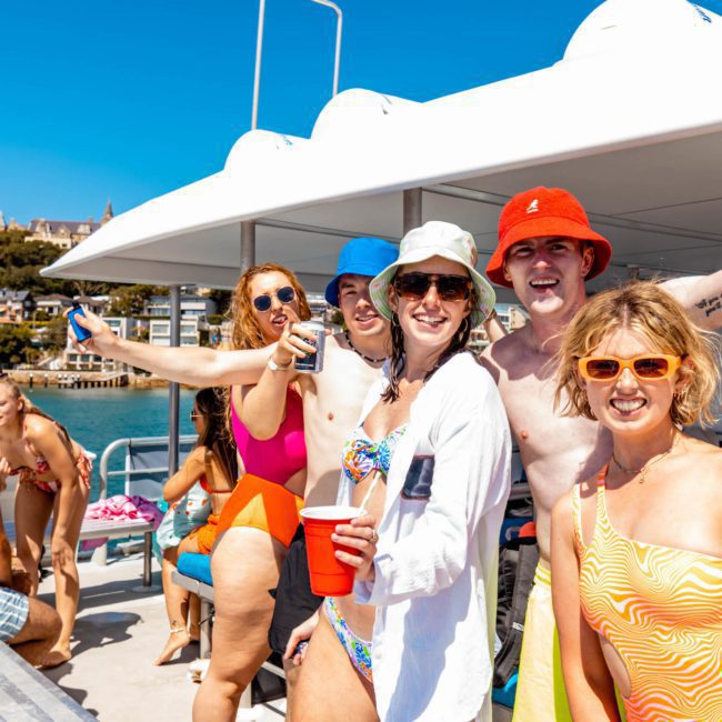 Group of people in swimsuits smiling and posing for a photo on a sunny day during a corporate boat event in Sydney. Some are holding drinks, and the background features a blue sky and distant shoreline.
