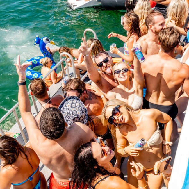 A group of people in swimwear are gathered on a boat, enjoying a sunny day on the water. Some are holding drinks while others swim nearby. This private yacht charter on Sydney Harbour is perfect for a fun and relaxing day with friends.