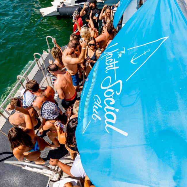 A group of people in swimwear are gathering on a luxury yacht hire Sydney near water under a blue canopy that reads "The Yacht Social Club." The scene suggests a private yacht charter Sydney Harbour, making for an exclusive social event or catamaran party.