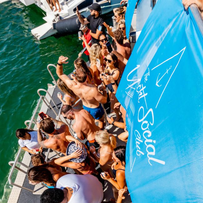 A group of people in swimwear gather on a boat deck by the water, with a banner that says "The Yacht Social Club." Some are dancing while others are sitting or standing, enjoying a sunny day. A DJ sets the mood, making it a perfect Sydney boat party hire experience.