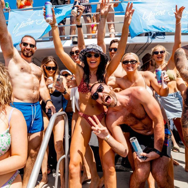 A group of people in swimwear are enjoying a Sydney boat party hire, holding drinks and posing excitedly for the camera. Some are raising their hands, and one person is making a peace sign.