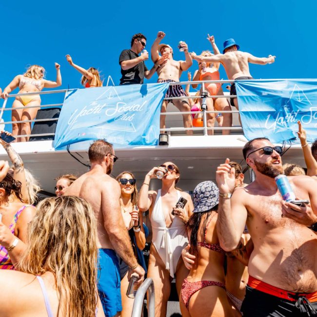A group of people in swimwear are partying on a boat under clear skies. Some are dancing on the upper deck while others are mingling on the lower deck. Blue banners are displayed along the railings, making it a perfect scene for a DJ boat hire Sydney event.