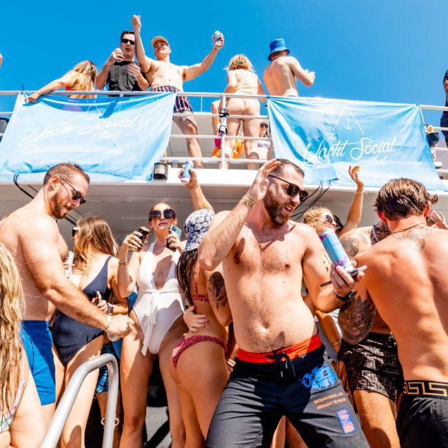 A group of people in swimwear are dancing and socializing on a crowded boat under a clear blue sky. A DJ boat hire Sydney keeps the party lively, with some guests holding drinks and banners displayed on the upper deck.