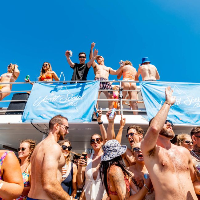 A large group of people are partying on a private yacht charter in Sydney Harbour under a clear blue sky. Some are on the upper deck, while others are below having drinks. Blue banners with "Yacht Social Club" are displayed on the boat, showcasing luxury yacht hire in Sydney.