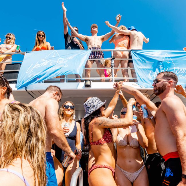 A group of people in swimsuits are gathered, socializing, and raising drinks on a multi-level boat deck under a clear, blue sky during a luxury yacht hire Sydney event.