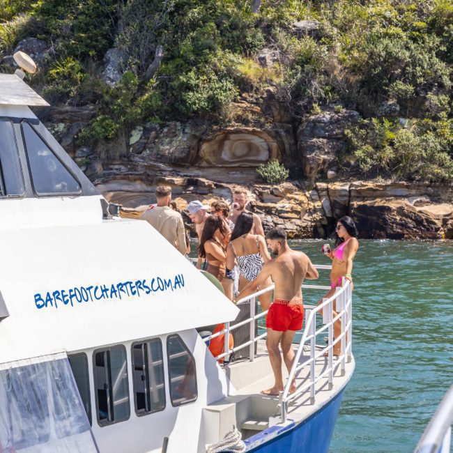 A group of people socializing on the deck of a luxury yacht from Barefoot Charters, with rocky terrain and trees in the background.