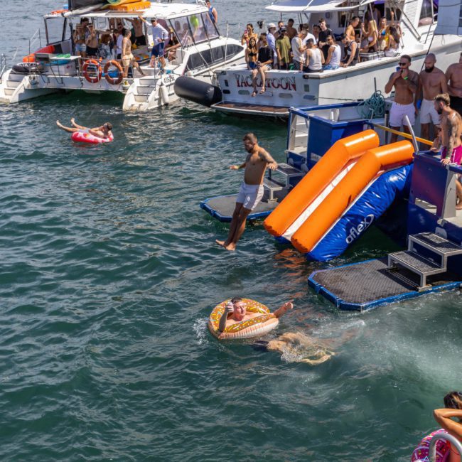 People enjoying a sunny day on a luxury yacht hire Sydney, with some using a slide to enter the water. A person floats in an inflatable ring while others swim or socialize nearby. Several boats, including private yacht charters in Sydney Harbour, are docked close together.