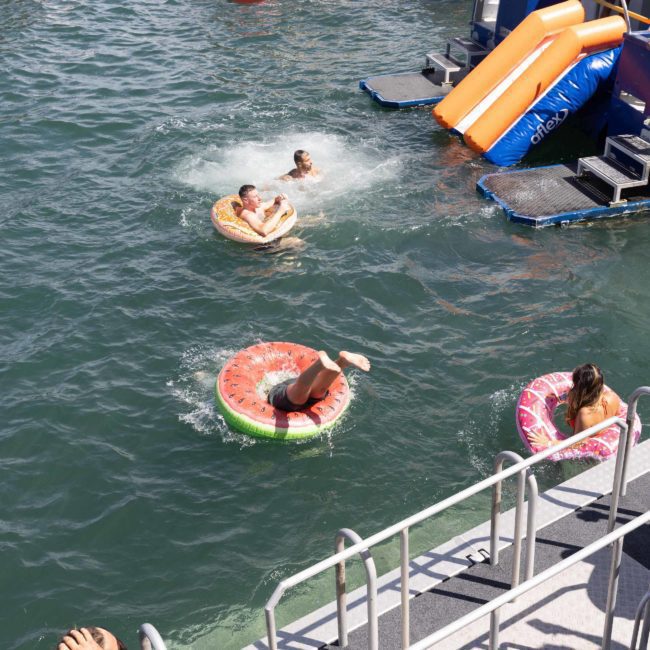 People enjoying water activities near boats. One person slides into the water while others float on inflatable tubes. A woman on the boat adjusts her hair. Watercraft and slides are visible in the background, creating a perfect scene for corporate boat events or a luxury yacht hire in Sydney.