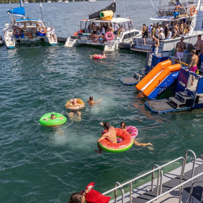 People enjoying a sunny day on the water with inflatable floats and slides near anchored boats, all part of a lively Catamaran party Sydney.