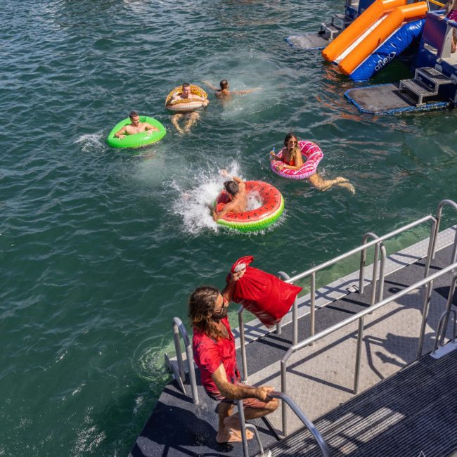 People in inflatable floats enjoy a water activity near a dock, with some entering the water from a slide. A person in a red shirt carries a red float up the steps. Experience similar fun with DJ boat hire Sydney or private yacht charter Sydney Harbour.
