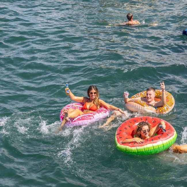 Three people are floating on inflatable rings shaped like a donut, a watermelon slice, and a pizza slice in a body of water during a Sydney boat party hire, holding drinks and smiling at the camera.