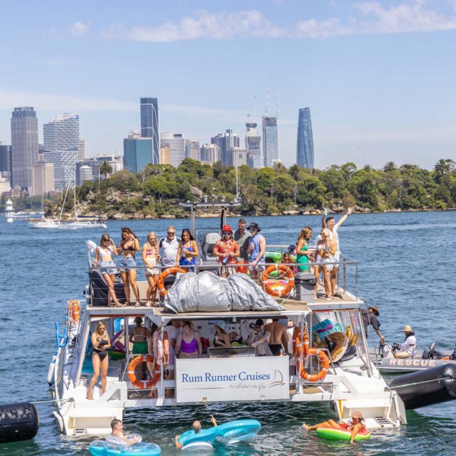 A group of people is gathered on a double-decker boat labeled "Rum Runner Cruises" on a sunny day. They are on the water with inflatable floats around them and a city skyline in the background, enjoying what looks like one of the best corporate boat events Sydney has to offer.