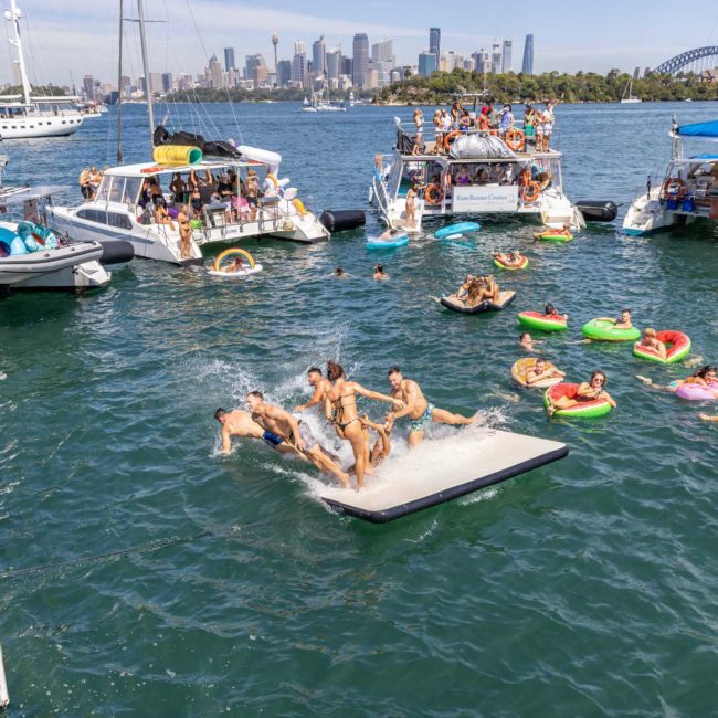 People enjoying a sunny day on boats and inflatables in a harbor, with a city skyline and a bridge in the background, celebrating with DJ boat hire Sydney.