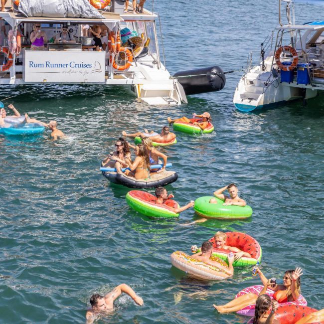 People in colorful inflatable pool floats enjoy the water near docked boats on a sunny day, with a cityscape and bridge in the background, setting the perfect scene for a Sydney boat party hire.