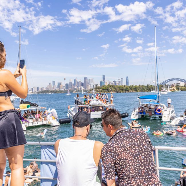 A group of people on a boat enjoy a sunny day, with some watching activities on other boats. A woman is taking a photo with her phone. The city skyline and a bridge are visible in the background, perfect for a catamaran party in Sydney or corporate boat events Sydney.