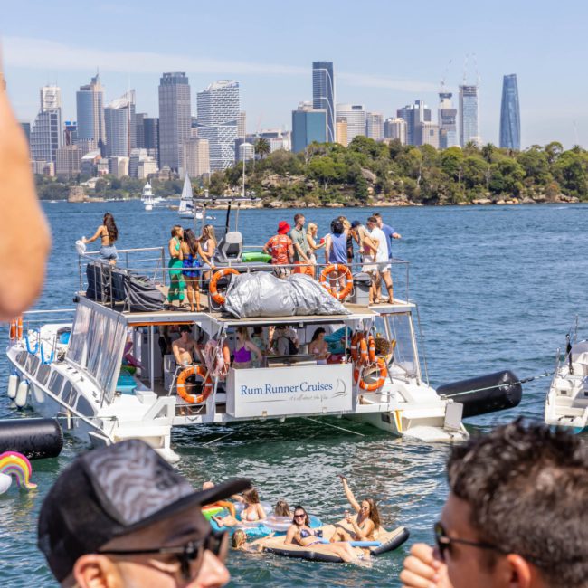 A group of people enjoy a sunny day on a luxury yacht labeled "Rum Runner Cruises" on the water, with a city skyline in the background and more people floating nearby on inflatables.