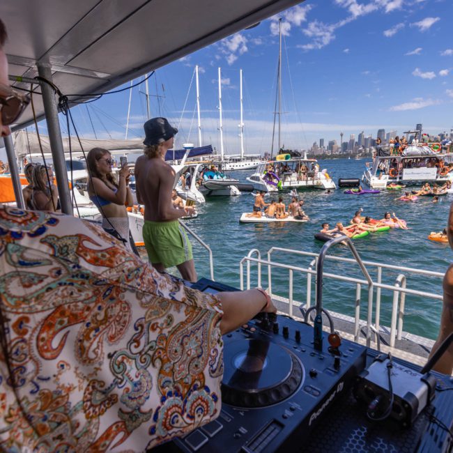 A DJ performs on a catamaran party in Sydney while people in swimsuits enjoy the water, floating on inflatables and gathered on nearby boats, with a city skyline in the background under a clear sky.