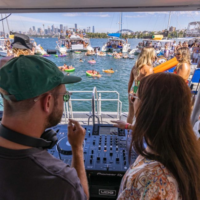 Two people stand at a DJ booth on a private yacht charter Sydney Harbour, overlooking a lively scene of people floating on inflatable rafts in the water. The city skyline is visible in the background.