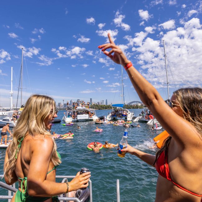 People on boats and in the water enjoy a sunny day; one woman in the foreground points upwards while holding a drink, as if inviting others to join her on a private yacht charter in Sydney Harbour.
