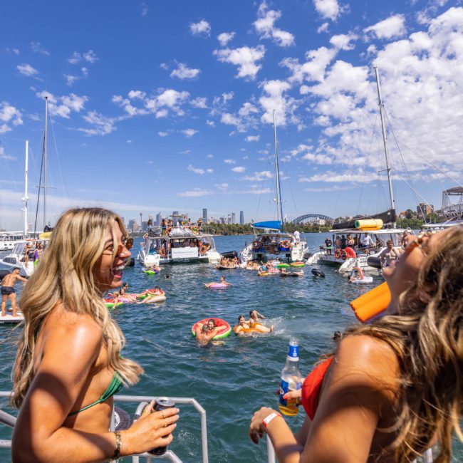 Two women on a dock smile and hold drinks, overlooking a lively scene of people on inflatable floats and boats in a marina under a partly cloudy sky. The setting hints at the fun atmosphere you can experience with DJ boat hire Sydney or even luxury yacht hire Sydney.