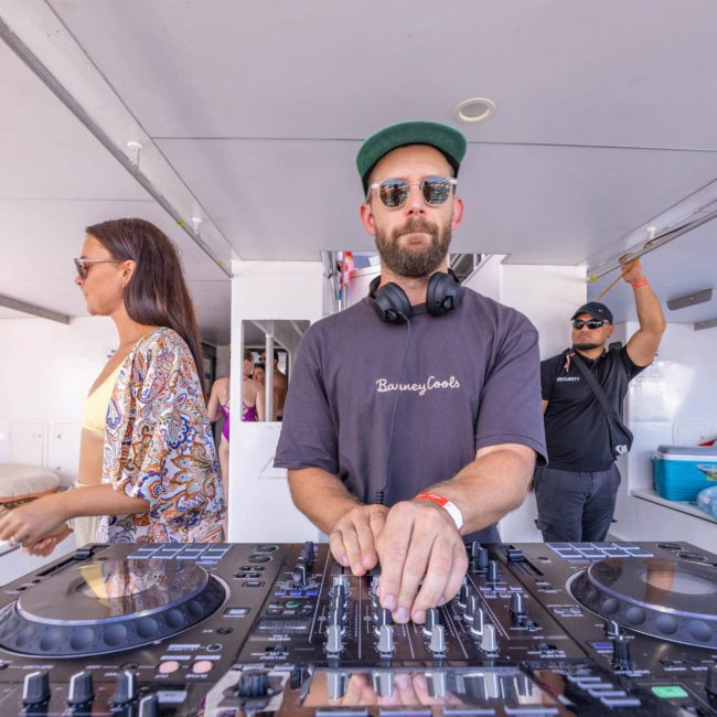 A man in sunglasses and headphones operates a Pioneer DJ mixer on a platform, with a woman and a security guard visible in the background. Perfect for those seeking luxury yacht hire Sydney or wanting to throw an unforgettable Sydney boat party hire experience.