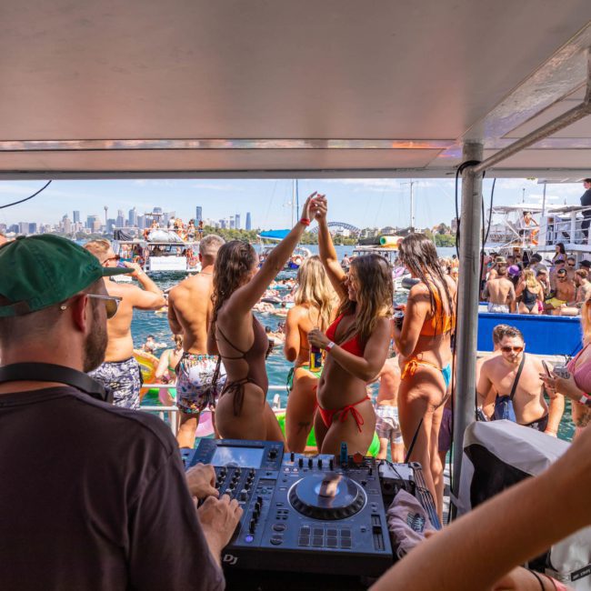 A DJ plays music on a luxury yacht hire Sydney while people in swimwear dance and enjoy a sunny day on the water, with the city skyline visible in the background.
