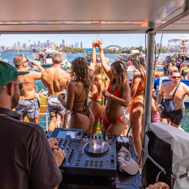 A DJ plays music on a boat filled with people in swimwear during a sunny day. Other boats and the city skyline are visible in the background, making it the perfect setting for a Sydney boat party hire.