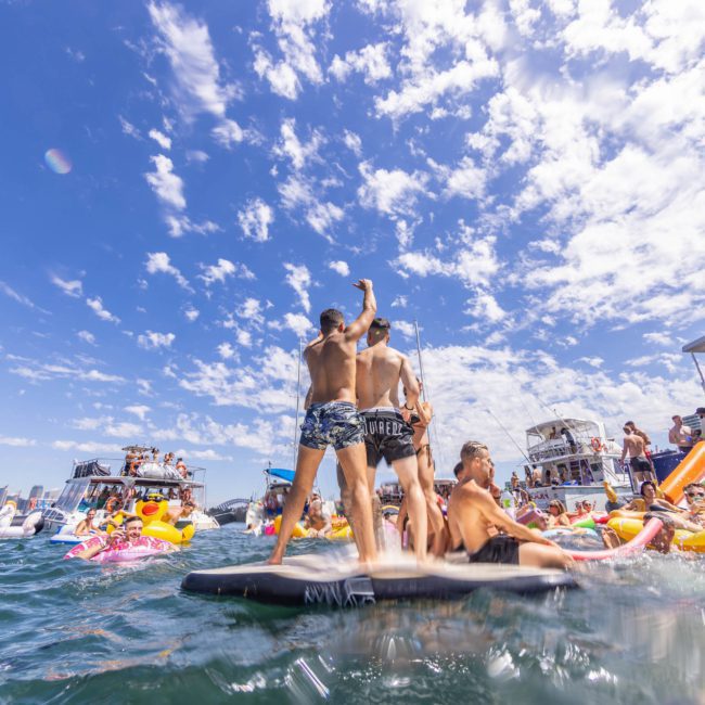 People are enjoying a sunny day on the water with inflatable floats and boats. Two individuals are standing on a float, with one raising an arm in the air. The sky is clear with scattered clouds, perfect for a Sydney boat party hire or DJ boat hire Sydney for a lively gathering.