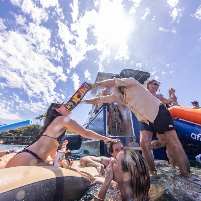 People enjoying a party on inflatable floats in the water, one person drinks from a novelty bottle while another pours. A boat and blue sky with scattered clouds are in the background, perfect for a Sydney boat party hire.