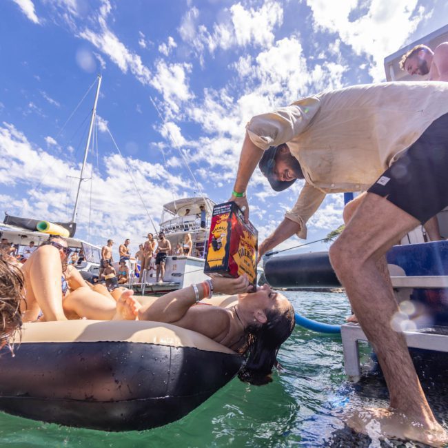 A person in a boat pours wine from a bag into the mouth of someone lounging on an inflatable float, surrounded by boats and people enjoying a partly cloudy sky. Luxury yacht hire Sydney adds an extra touch to this idyllic scene.