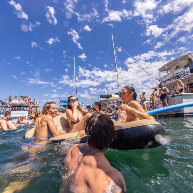 People on floatation devices and boats enjoying a sunny day on the water, with some carrying drinks and swimming. Sailboats are docked in the background under a partly cloudy sky, ideal for a Catamaran party Sydney.