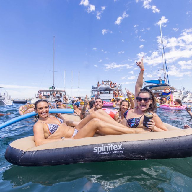A group of people are enjoying a sunny day on the water, using inflatable floats. Some are on paddleboards, while others are on boats in the background. The sky is clear with a few scattered clouds, making it a perfect day for a private yacht charter in Sydney Harbour.