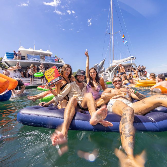 Four people are lounging on an inflatable raft in the water, surrounded by other rafts and boats, beneath bright sunlight and clear skies. It’s the perfect day for a Catamaran party Sydney style.