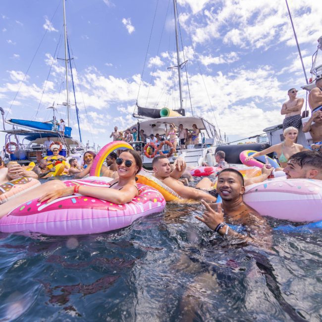 A group of people are gathered on inflatable pool floats in the water near docked boats, enjoying a sunny day with a partly cloudy sky above. It looks like the perfect setup for a catamaran party Sydney style, bringing together fun and relaxation on the water.