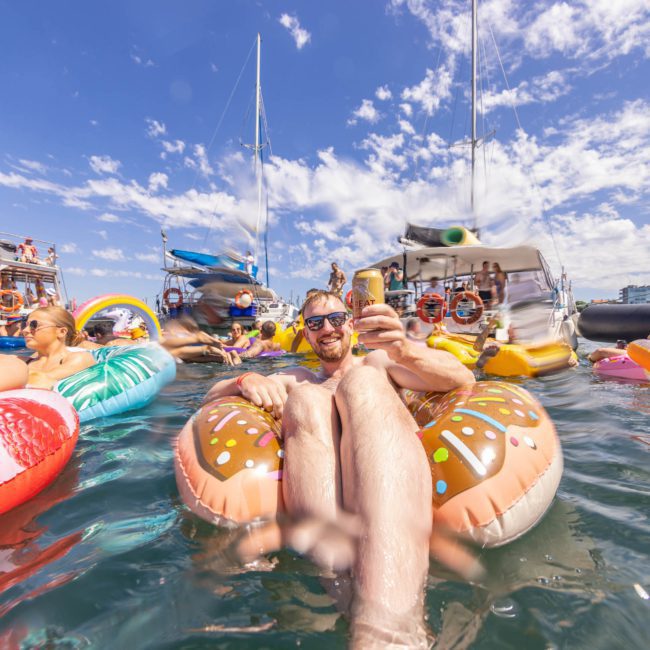 A man relaxes on a donut-shaped float in the water, surrounded by others on various inflatables, with boats and clear skies in the background—a perfect ambiance for a Sydney boat party hire.