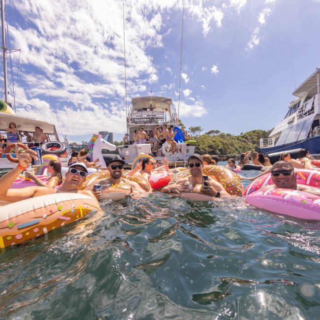 A group of people enjoy floating on inflatable rings in the water near docked yachts on a sunny day, celebrating a lively Sydney boat party hire.