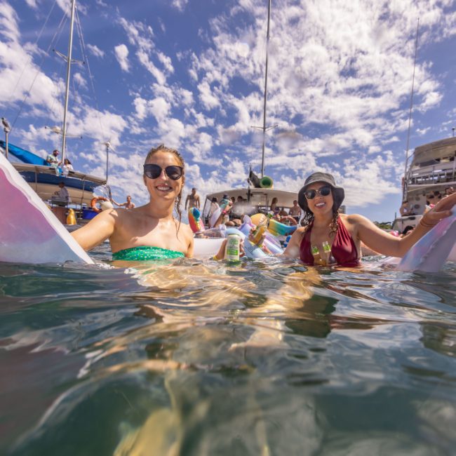 Two people relax on floaties in the water near luxury yacht hire Sydney boats, holding drinks and smiling under a partly cloudy sky.