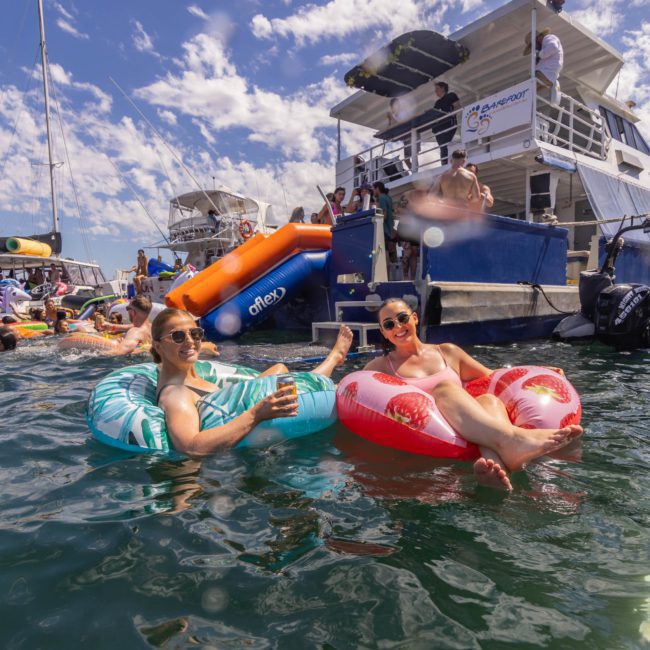 Two people floating on inflatable pool rings in the water with a boat and other people having fun in the background, epitomizing a perfect day of luxury yacht hire Sydney.
