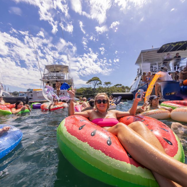 People on inflatable floats enjoy a sunny day in the water near boats, with a private yacht charter on Sydney Harbour against a backdrop of clear sky and clouds.