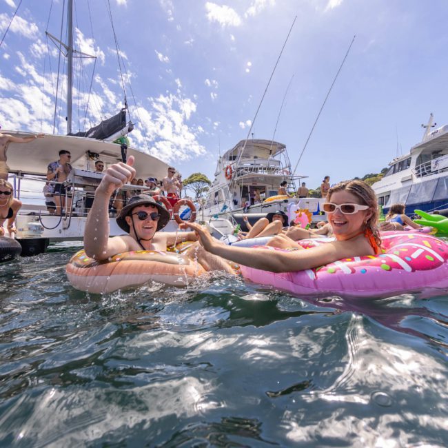 People relaxing on inflatable floats in the water surrounded by boats under a clear sky. Some are interacting and enjoying the sunny day, with luxury yacht hire Sydney enhancing the perfect atmosphere.