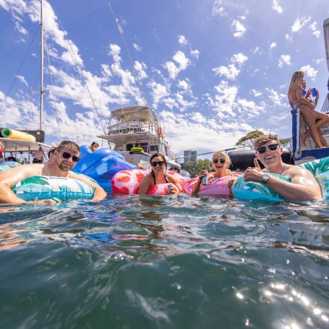 A group of people on inflatable floaties enjoying a sunny day in the water, with boats and other people in the background, creating the perfect setting for a Sydney boat party hire.