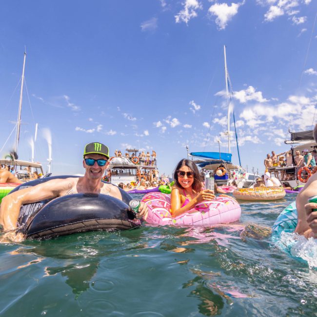 People are floating on inflatable rafts and enjoying drinks in the water near anchored boats on a sunny day, with music from a DJ boat hire Sydney adding to the festive atmosphere.