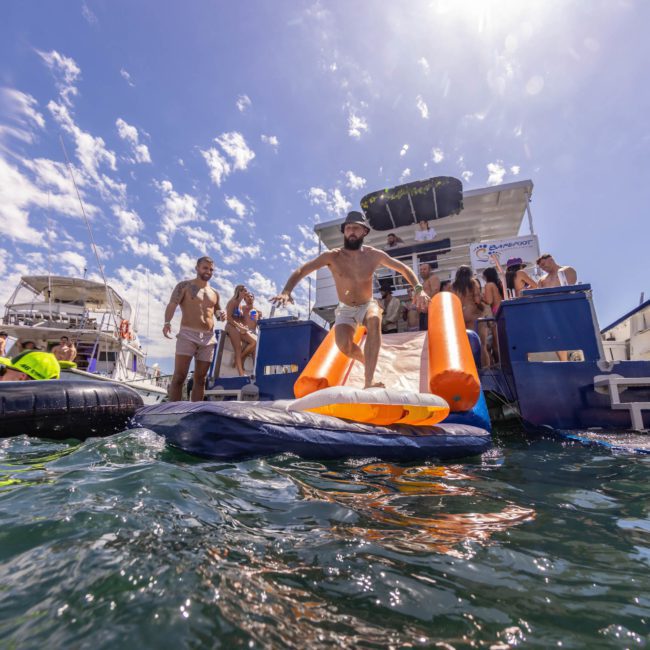 A man prepares to slide into the water from a boat slide, joined by others on a sunny day. Several boats, including a luxury yacht hire Sydney, are in the background.