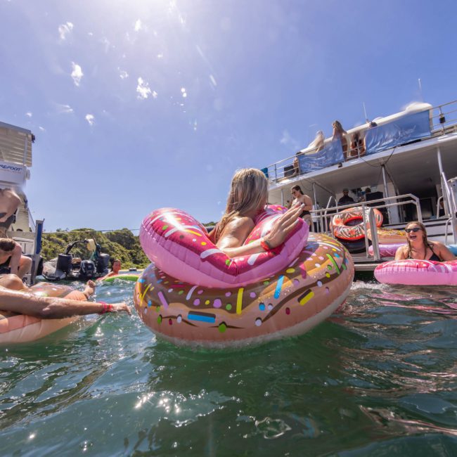 People are enjoying a sunny day on a lake, floating on inflatable pool toys near boats. A person in the foreground is on a donut-shaped inflatable ring, soaking up the atmosphere of luxury yacht hire Sydney.